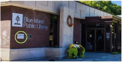 The image shows the front entrance of the Blue Island Public Library (located at 2433 York St., Blue Island, IL). The yellow circle highlights the library's external book drop box (also called the return/drop box). This is the small rectangular metal slot mounted on the brick wall to the left of the main entrance doors. It is available 24/7 for returning library materials, even when the building is closed.